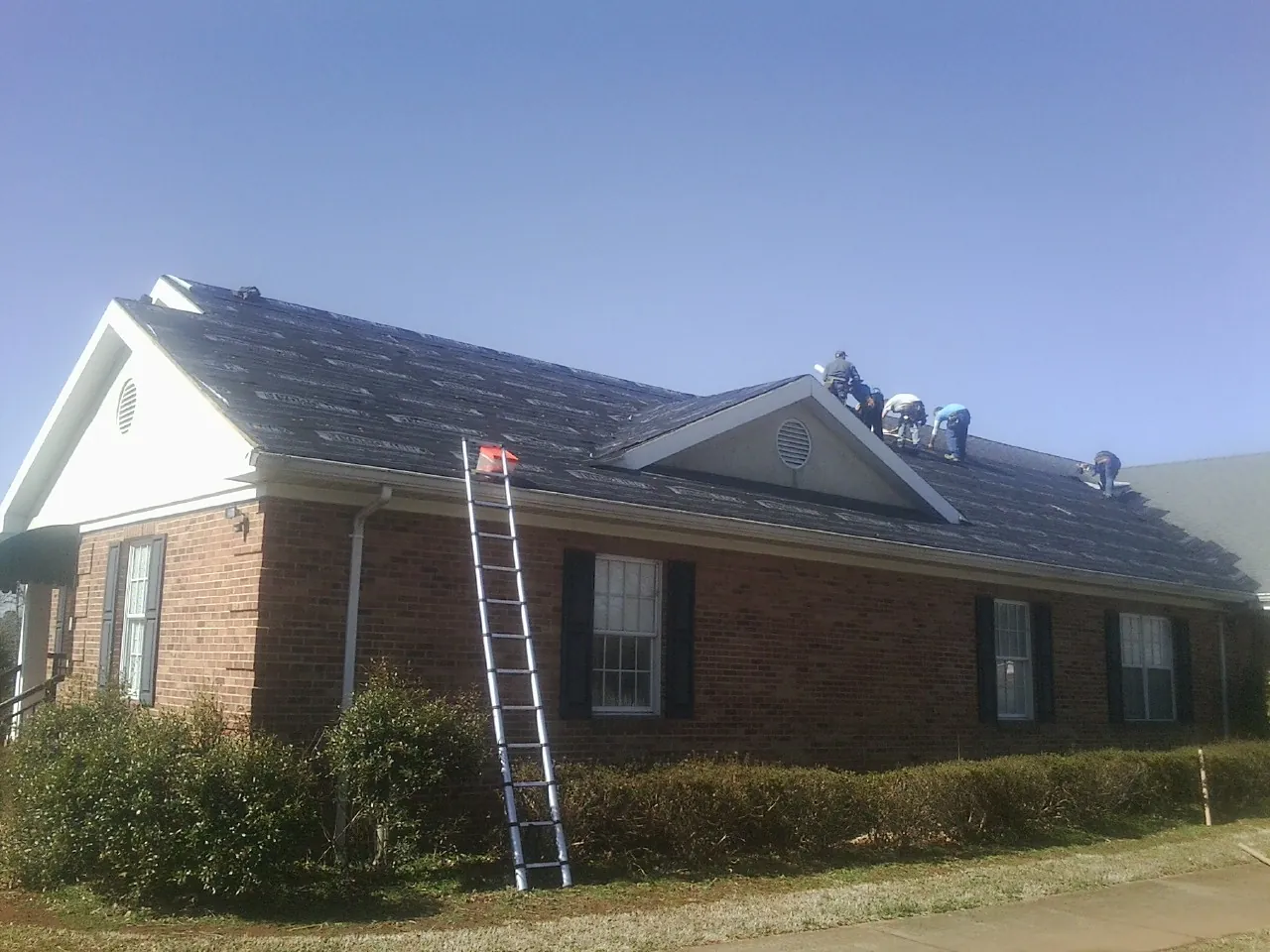 Crew tearing shingles off a brick ranch roof with a ladder set — Emergency Roof Repair work by our crew in Pleasant Valley